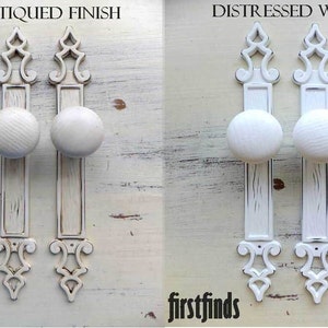 May include: Two white door knobs with ornate metal plates in two finishes: antiqued and distressed white. The knobs are round and have a raised design. The plates are rectangular with a decorative scroll design at the top.