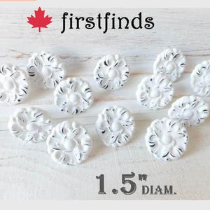 May include: A collection of twelve white circular knobs with a distressed black finish, each featuring a raised floral pattern. The knobs are arranged in a group and measure 3.81 cm in diameter as indicated by the text at the bottom.