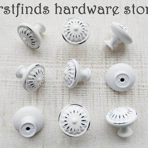 May include: White cabinet knobs with a distressed black trim. The knobs have a floral design on the face and are arranged on a light wood surface. The text "firstfinds hardware store" is at the top.