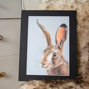 May include: A framed art print featuring a detailed pencil drawing of a hare. The hare has brown fur, long ears, and a focused gaze. The artwork is set against a light gray background and framed in a black mat and frame.