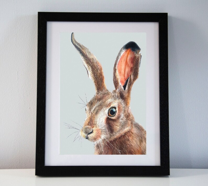 May include: A framed art print featuring a detailed illustration of a brown hare. The hare has long ears, detailed fur, and a focused gaze. The artwork is set against a light gray background and framed in a black frame, suitable for home decor.