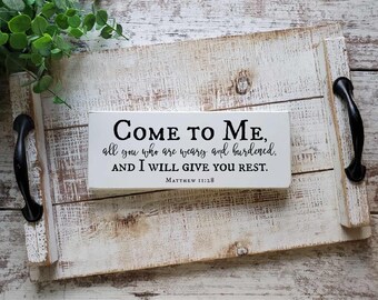Puede incluir: Un letrero rectangular de madera blanca con texto negro que dice Come to Me, all you who are weary and burdened, AND I WILL GIVE YOU REST. Matthew 11:28, sobre una bandeja de madera blanca desgastada con asas de metal negro.
