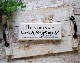 May include: A rectangular white wooden sign with black text that reads Be strong & Courageous Joshua 1:9 For the Lord your God is with you wherever you go, resting on a distressed white wooden tray with black metal handles.