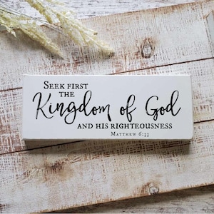 May include: White wooden sign with black text that reads "Seek first the Kingdom of God and His Righteousness Matthew 6:33". The sign is sitting on a white wooden tray with black handles.