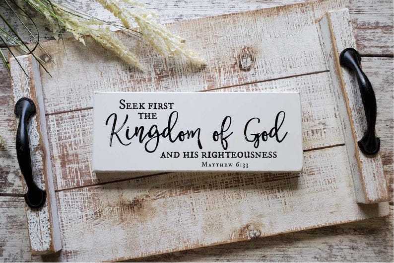May include: White wooden sign with black text that reads "Seek first the Kingdom of God and His Righteousness Matthew 6:33". The sign is sitting on a white wooden tray with black handles.
