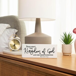 May include: White wooden sign with black text that reads "Seek first the Kingdom of God and His righteousness Matthew 6:33". The sign is sitting on a wooden nightstand with a white alarm clock and a pink vase with pink flowers.