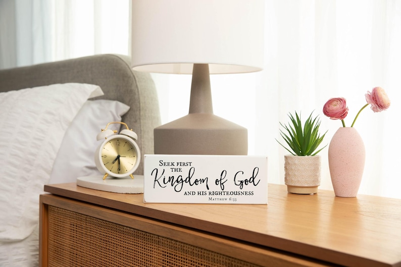 May include: White wooden sign with black text that reads "Seek first the Kingdom of God and His righteousness Matthew 6:33". The sign is sitting on a wooden nightstand with a white alarm clock and a pink vase with pink flowers.