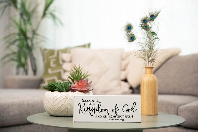 May include: White wooden sign with the text "Seek first the Kingdom of God and His righteousness Matthew 6:33". The sign is sitting on a table with a potted succulent plant and a vase with peacock feathers.