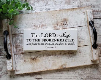 Puede incluir: Un letrero rectangular de madera blanca con texto en negro que dice: The LORD is close TO THE BROKENHEARTED AND saves those who are crushed in spirit. Psalm 34:18. El letrero está sobre una bandeja de madera blanca desgastada con asas de metal negro.