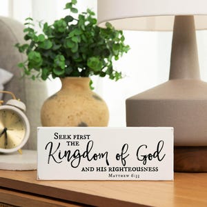 May include: White wooden sign with black text that reads "Seek first the Kingdom of God and His righteousness Matthew 6:33". The sign is sitting on a wooden surface next to a white alarm clock and a vase with green plants.