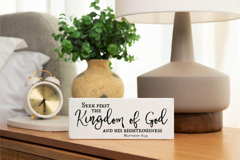 May include: White wooden sign with black text that reads "Seek first the Kingdom of God and His righteousness Matthew 6:33". The sign is sitting on a wooden surface next to a white alarm clock and a vase with green plants.