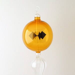 May include: A glass Christmas ornament with a clear, teardrop-shaped bottom and a round, amber-colored top. The top has a small, clear glass loop for hanging and a black, geometric design inside.