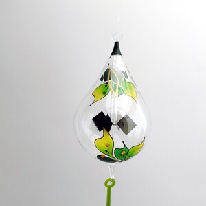 May include: A clear glass teardrop ornament with a green and yellow painted butterfly design. The ornament has a black metal top and a green glass drop hanging from the bottom.