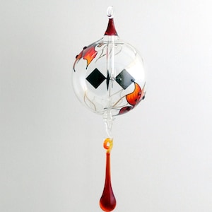May include: A glass ornament with a red and orange design of leaves and a small black propeller inside. The ornament is suspended from a string and has a red glass teardrop hanging from the bottom.