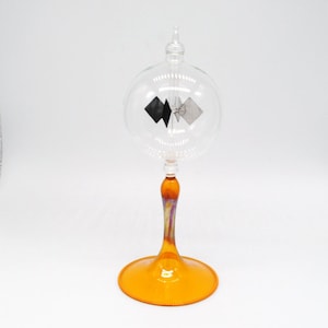 May include: A glass sphere with a small, rotating, black and silver object inside, mounted on a clear glass stem with an amber colored base.