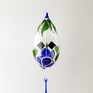 May include: A clear glass ornament with a blue and green floral design. The teardrop-shaped ornament has a blue top and a dangling blue teardrop. Green leaves and blue flowers decorate the glass.