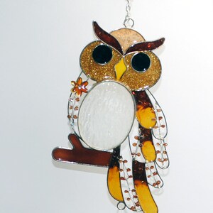 May include: A stained glass owl suncatcher with a brown branch, gold glitter accents, and a clear glass teardrop-shaped ornament with orange floral designs. The owl has a white belly and is hanging from a silver chain.