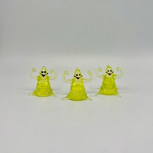 May include: Three small glass figurines of ghosts with arms raised in a flexing pose. The ghosts are yellow and translucent with black eyes and a black smile.