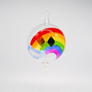 May include: A glass ornament with a rainbow painted on it. The rainbow is made up of the colors red, orange, yellow, green, blue, indigo, and violet. There is a small blue cloud painted on the bottom of the ornament.