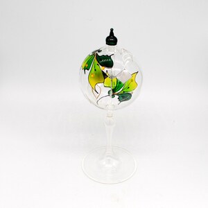 May include: A clear glass bottle stopper with a green and yellow butterfly design. The bottle stopper is on a clear glass stand with a round base.