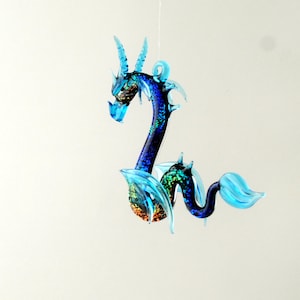 May include: A blue glass dragon ornament with iridescent glitter accents. The dragon has a long tail and wings, and is hanging from a string.