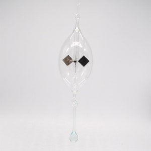 May include: A clear glass ornament with a small, dark-colored propeller inside. The ornament is suspended from a thin string and has a teardrop-shaped bottom.