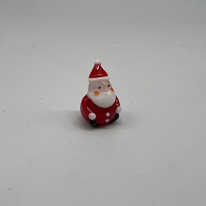 May include: A small glass figurine of Santa Claus wearing a red suit with white trim and a white beard. He has a red hat with white trim and a white pom-pom on top.