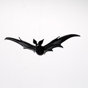 May include: A black bat shaped decoration hanging from a string. The bat has a rounded body and pointed wings.