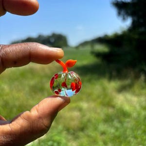 以下が含まれることがあります： カボチャや果物の形をした小さな透明なガラスの置物で、内部に赤い斑点があり、鮮やかなオレンジ色の茎が付いています。ガラスのオブジェは半透明で、丸みを帯びた本体を光が透過し、鮮やかな赤い斑点が強調されています。