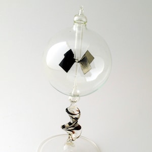 May include: A glass radiometer with a clear glass bulb and a silver base. The bulb contains a four-blade rotor with black and silver vanes that spin when exposed to light.