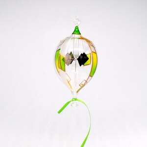 May include: A clear glass ornament with green, yellow, and white accents. The ornament has a bulbous shape with a green top and a green ribbon. Inside are black and silver diamond shapes. The ornament hangs from a thin string.