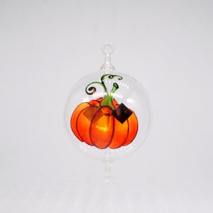 May include: A glass ornament with a painted pumpkin inside. The pumpkin is orange with green leaves and a stem. The ornament is clear glass and has a silver hanger.