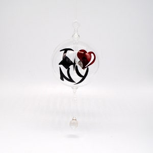May include: Clear glass ornament with the text "I NY" in black script and a red heart. The ornament has a loop at the top for hanging and a decorative glass element at the bottom. The ornament is suspended against a white background.