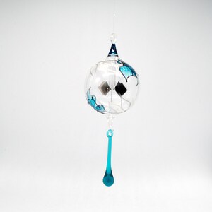 May include: A clear glass wind chime with a spherical body, adorned with blue leaf-like designs and silver accents. The chime features a teardrop-shaped pendant and a dark blue top, suspended by a thin, clear string. The overall design is delicate and elegant.