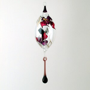 May include: A clear glass ornament with a teardrop shape, featuring a dark purple and red floral design. The ornament has a dark purple top and a matching teardrop-shaped dangle. The background is plain white.