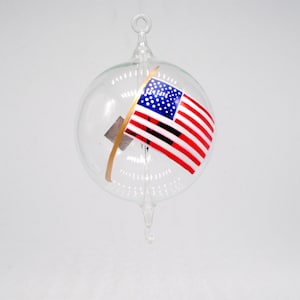 May include: A glass ornament with a red, white, and blue American flag inside. The flag is painted on the inside of the glass sphere and is slightly tilted.