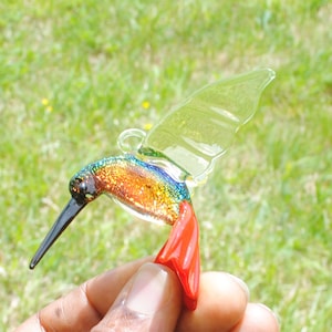 May include: A glass hummingbird ornament with iridescent wings and a red tail. The hummingbird is in flight and has a black beak.