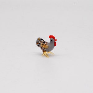 May include: A small glass rooster figurine with a red comb and speckled brown and grey feathers.
