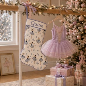 Könnte beinhalten: Ein cremefarbener Weihnachtsstrumpf mit einem wiederkehrenden Muster aus Ballerinas in blau-weißen Tutus, auf dem der Name Quinn in Blau gestickt ist. Daneben hängt ein lavendelfarbener Ballett-Trikot mit mehrlagigem Tüllrock und Goldbesatz.