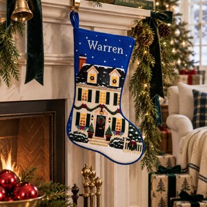 Può includere: Una calza natalizia blu con un'illustrazione a punto croce di una casa a due piani con finestre illuminate, una ghirlanda sulla porta d'ingresso e terreno innevato. Il nome Warren è ricamato in bianco nella parte superiore della calza.