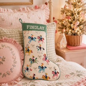 Puede incluir: Un calcetín navideño de color crema con un puño verde que lleva el nombre FINDLAY bordado en blanco. El cuerpo del calcetín muestra un patrón repetitivo de jinetes coloridos a caballo y pequeños lazos en varios tonos de rosa y azul.