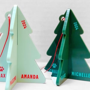 May include: Two green and blue Christmas tree ornaments with red string and the year 2024 printed on them. The ornaments are personalized with the names Max, Amanda, Michelle, and Chill.