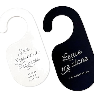 May include: Two door hangers with a hook top. The white hanger features black text reading Shh.. Session in Progress and PLEASE DO NOT DISTURB. The black hanger features white text reading Leave me alone. and I'M MEDITATING in a script font.