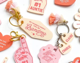 May include: A collection of pink and peach-colored keychains, bookmarks, and decorative items with phrases like "Best Grandma Ever" and "#1 Auntie." Some items include tassels and gold-colored hardware. The items are designed with a loving theme.