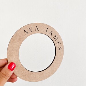 May include: A circular, light brown wooden cutout with the name "AVA JAMES" engraved in black, held by a hand with red nail polish. The cutout has a circular opening in the center.