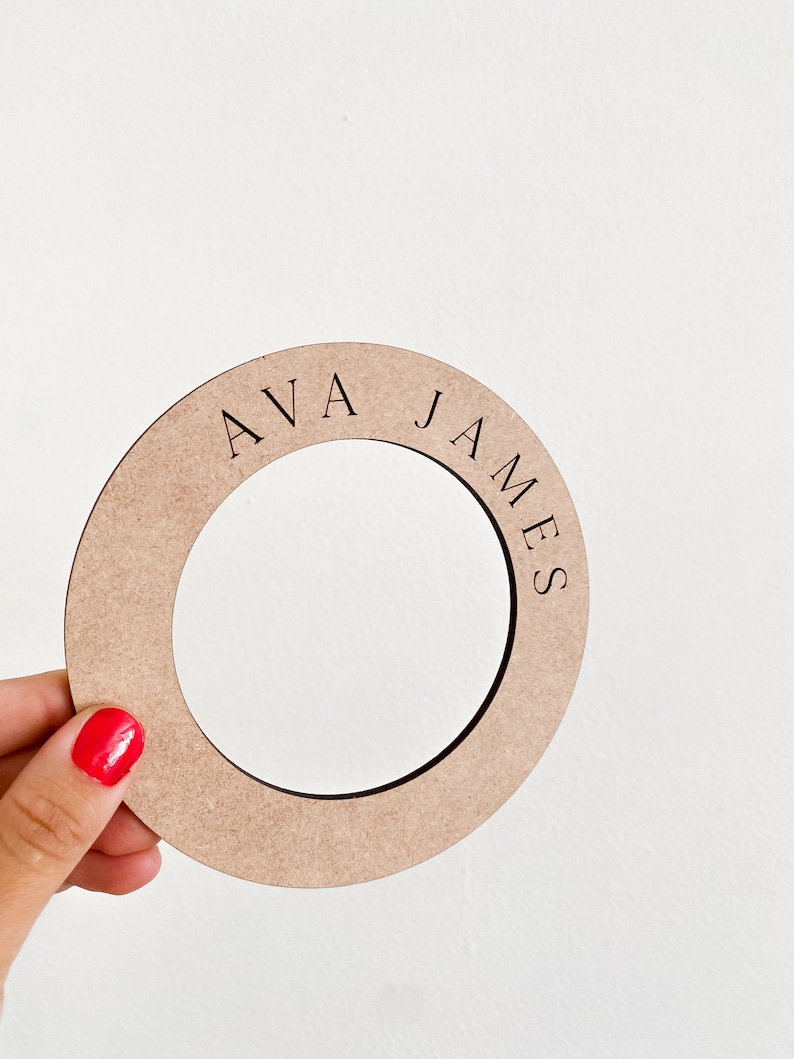 May include: A circular, light brown wooden cutout with the name "AVA JAMES" engraved in black, held by a hand with red nail polish. The cutout has a circular opening in the center.