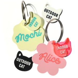 May include: Five colorful cat ID tags with key rings. Two tags are yellow with the text "Indoor Cat" and "Mochi". One tag is pink with the text "Alice" and "Indoor Cat". One tag is white with the text "Outdoor Cat". One tag is black with the text "Outdoor Cat".