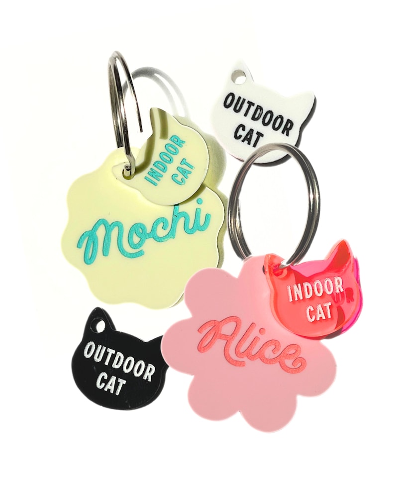 May include: Five colorful cat ID tags with key rings. Two tags are yellow with the text "Indoor Cat" and "Mochi". One tag is pink with the text "Alice" and "Indoor Cat". One tag is white with the text "Outdoor Cat". One tag is black with the text "Outdoor Cat".