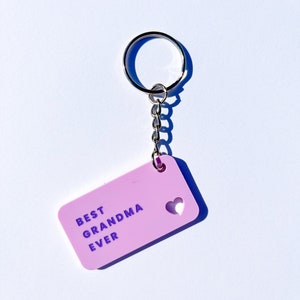 May include: A pink keychain with a silver key ring and chain. The keychain has a heart-shaped cutout and the text "BEST GRANDMA EVER" in purple lettering.
