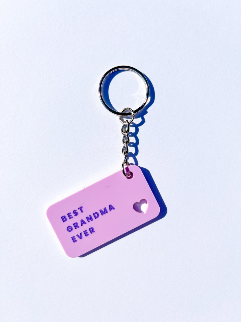 May include: A pink keychain with a silver key ring and chain. The keychain has a heart-shaped cutout and the text "BEST GRANDMA EVER" in purple lettering.